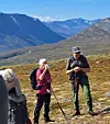 To turgåere med staver står i et steinete fjellandskap med fjell i bakgrunnen.