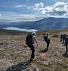 En gruppe turgåere krysser et steinete fjellplatå med innsjø og fjell i bakgrunnen.