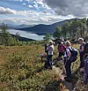 Turgåere på en fjellsti med utsikt over innsjø, skog og fjell under en skyfull himmel.