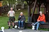 Tre personer ved en trekonstruksjon i en grønn hage, med en rullestol og benker.