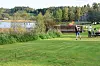 Grønn gresslette med mennesker ved en treplattform, vann og skog i bakgrunnen. En dame kaster hestesko