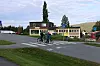 To syklister krysser en vei foran bygninger og grønt landskap.