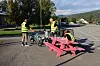 Tre personer i refleksvester og en gutt på en stol ved sykler og rosa benk på en parkeringsplass.