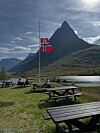 Norsk flagg på halv steng foran et spisst fjell ved en innsjø, med rastebord i forgrunnen.
