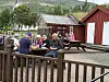En gruppe mennesker sitter ved et trebord utenfor en gårdskafé i landlige omgivelser.