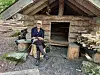 Person sitter foran en gammel tømmerhytte med vedstabler og steinete grunn.