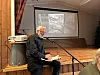 Foredragsholder ved mikrofon foran et lerret med et svart-hvitt bilde i en trepanelt sal.