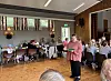 Eldre mennesker samles rundt bord i et lyst lokale, med en kvinne i rosa jakke stående i midten.