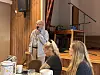 Taler med mikrofon foran tre sittende personer i et lokale med piano.