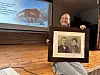 Kvinne holder et innrammet historisk fotografi foran en projisert presentasjon på en scene.