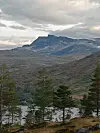 Fjell med innsjø og furutrær under en overskyet himmel.