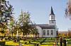 Hvit kirke med høyt spir, og gravstøtter og trær på en kirkegård.