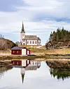 Hvit kirke og rødt uthus speiler seg i et stille vann/sjø/fjord under en skyet himmel.