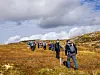 Turgåere går i rekke over en åpen fjellvidde med skyer og sollys i bakgrunnen.