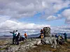 Turgåere ved en steinvarde på en steinete fjelltopp med fjellutsikt og skyet himmel.