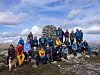 Turgåere i fargerike klær står samlet ved en steinvarde på en fjelltopp.