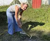 Kvinne bruker et hageredskap på bakken i en solfylt hage ved en rød bod.