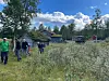 En gruppe mennesker går gjennom en frodig eng mot hus og skog under en blå himmel.