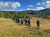 Turgåere går på rekke gjennom en åpen eng med skog og fjell i bakgrunnen.