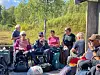 Gruppe syklister med hjelmer og bagasje tar pause utendørs ved skogkanten.
