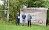 Tre personer står foran en trevegg med malte symboler i grønne omgivelser.