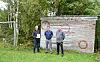 Tre personer står foran en trevegg med malte symboler i grønne omgivelser.