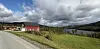 Rødt hytte og et gammelt fjøs ved en grusvei med grønne jorder, innsjø og skogkledde åser.