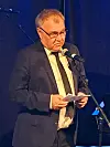 Mann i dress taler i mikrofon og leser fra notater på en blåbelyst scene.