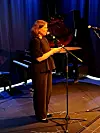 Ei dame som snakker i mikrofon på en scene med flygel og scenebelysning.