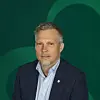 Mann i mørk dressjakke foran en grønn bakgrunn med buemotiv.
