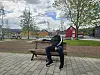 Mann sitter på en benk på et torg med trær og fargerike trehus i bakgrunnen.