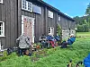 Gruppe mennesker sitter og står på gresset utenfor en eldre trebygning.
