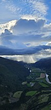 Dramatiske skyer over en grønn fjelldal med elv, åkre og solstråler.