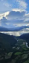 Dramatiske skyer over en grønn fjelldal med elv, åkre og solstråler.