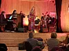 Vokalist synger foran et band på en opplyst scene, med publikum sittende i forgrunnen.