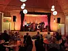 Musikere på en rødt opplyst scene foran et sittende publikum i en konsertsal.