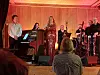 Vokalist og musikere opptrer på en liten trescene foran et sittende publikum.