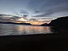 Solnedgang over en fjord med mørke fjell, speilblankt vann og strand i forgrunnen.