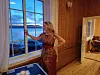 Kvinne ved et stort vindu i et trehus, med vannutsikt og et åpent rom bak seg.