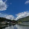 Liten fjordbygd ved rolig vann, omgitt av grønne fjell og skyer på blå himmel.