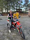 Motocrossfører med hjelm ved siden av en rød sykkel med nummer 496 på en grusplass.