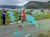 Deltakere løper gjennom turkis fargepulver ved et fjellomkranset vann.