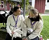 To kvinner med navneskilt sitter på gresset og snakker utenfor en trebygning.