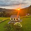Hvitt hus i grønne omgivelser med blomster, fjell og solnedgang.