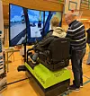 En ung mann prøver en gravemaskinsimulator med tre skjermer. En mann står på siden og instruerer.