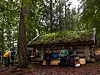 Turgåere står foran en liten mosegrodd tømmerhytte i skogen.