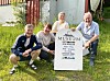 Fire personer står og sitter ved et hvitt museumsskilt på en gressplen.