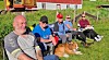 Gruppe personer sitter på gresset med en hund foran en rød bygning.