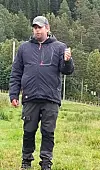 Mann i mørke friluftsklær står på en gresslette foran tett barskog.