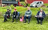 Fire personer sitter på stoler i en gresskledd utendørs setting med kjøretøy bak seg.